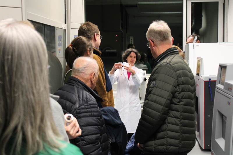 Das Bild zeigt die Abteilungsleiterin Kerstin Schöberl bei einer Laborführung im Bereich Elementanalytik. In der Hand hält sie ein dickwandiges Glasgefäß, in dem die Proben aufgeschlossen werden. Im Vordergrund sieht man Besucherinnen und Besucher.